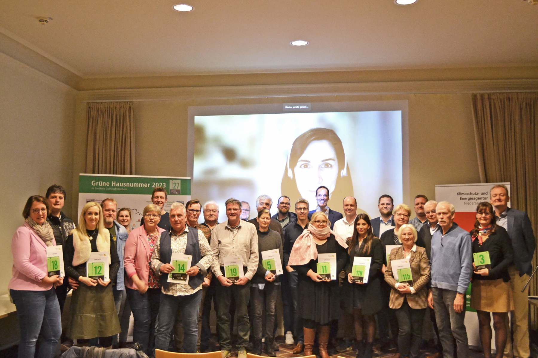 Erster Kreisrat Dr. Michael Kiehl (hintere Reihe Mitte) und Carmen Dittmer (KEAN, online zugeschaltet) konnten 21 grüne Hausnummernschilder überreichen. Bild: Landkreis Grafschaft Bentheim.